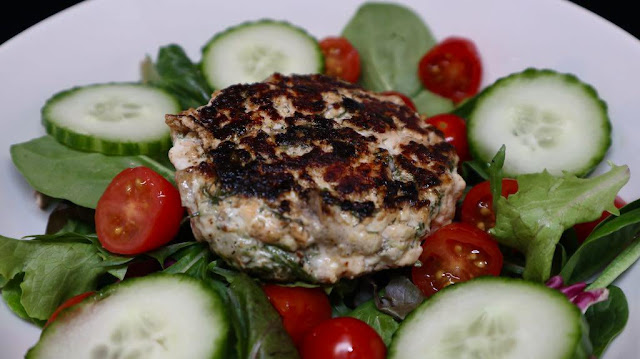 white plate, salmon burger on salad with cucumbers and tomatoes