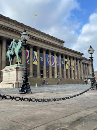 St George's Hall in Liverpool. The sky is blue, there's a Ukrainian flag flying from a mast on the roof, the 2023 Eurovision logo and colour are on banners coming down from the eaves. They're heart shape-stripes of blue, yellow, pink, and black.