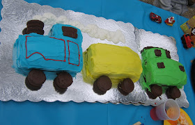 A train cake, sideways view, with cupcake wheels on a Engine with a double cupcake smokestack, a box car, and a caboose.   There whip cream "smoke"  and it's sitting on two overlapped silver cardboard trays, on top of a blue plastic tablecloth.   There are red decorative details on the Engine.