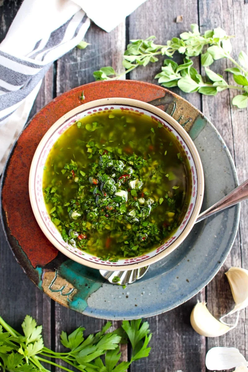 Top view of Chimichurri Sauce in a tan bowl on a ceramic multi-colored plate placed on a rustic wood background.