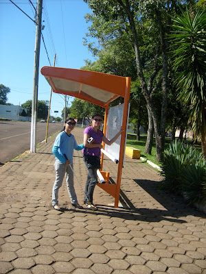 Uma parada de ônibus fotografada lateralmente, ela fica bem no meio da calçada, ao lado estão Tania e Alessandra em pé.