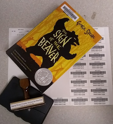 Stack of books, only the top book's cover visible, atop a sheet of barcode labels with a school address stamp and stamp pad next to them. The top book's title identifies it as 'The Sign of the Beaver' by Elizabeth George Speare. The image on the cover depicts the dark outline of a large creature rearing up on its hind legs against a yellow background