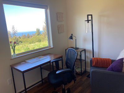 Office view, showing a small desk pulled to a window--vase with flowers on the sill. Window view shows Alaskan landscape at mid-day, water and pines. Office decor includes roller chair, lamps, and purple comfy chair..