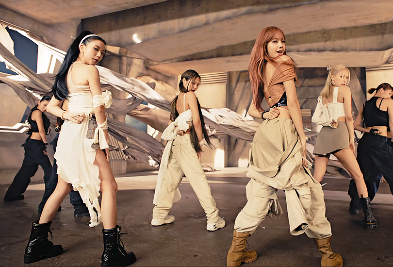 A screenshot from Blackpink’s music video for “Pink Venom”. Featuring all 4 members stood with their backs to the camera, in cream and white ensembles, in a stone room as beige curtains blow dramatically in the background.