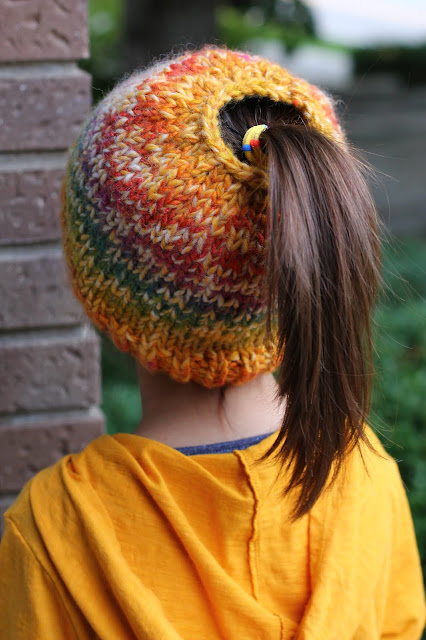 From the back of a child with light brown hair in a pony tail that comes through the hole on the top of a rainbow and yellow knit beanie. Child is wearing a hooded top that's sunshine yellow.