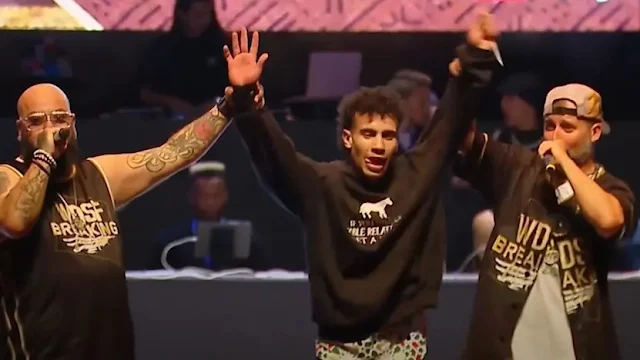 B-boy Billy, com casaco preto, tem as mãos levantadas pelos apresentadores indicando sua vitória. Ele é moreno e está com a língua para fora