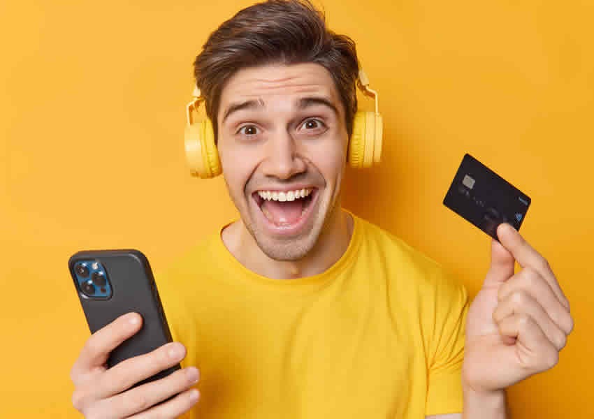Imagem mostra um homem sorrindo vestido com uma camisa amarela e fones de ouvido amarelo em uma das mãos um smartphone na outra mão um cartão de crédito.