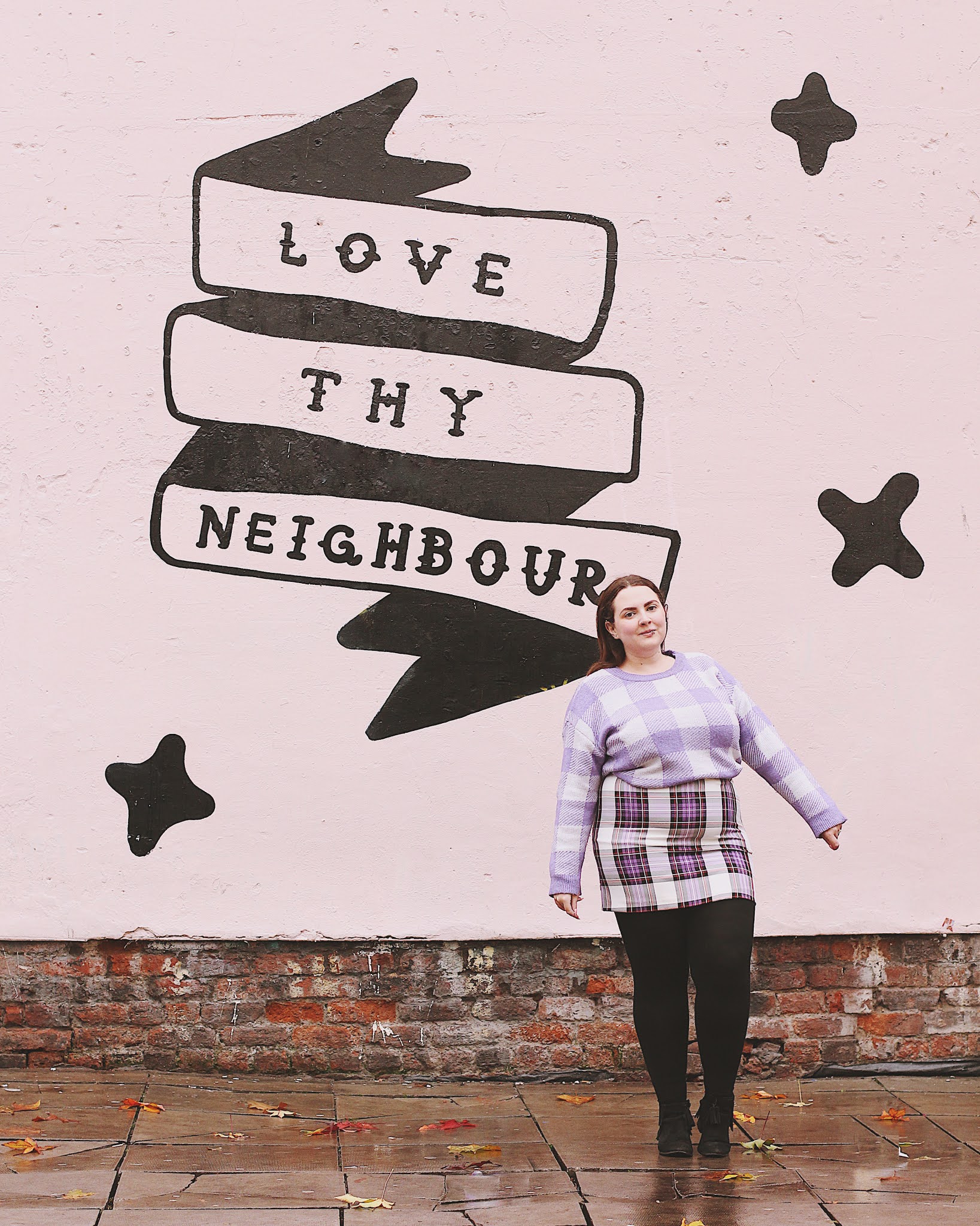 Becky stands in a purple checked jumper and skirt, in front of a pink wall with 'love thy neighbour' painted on as a mural