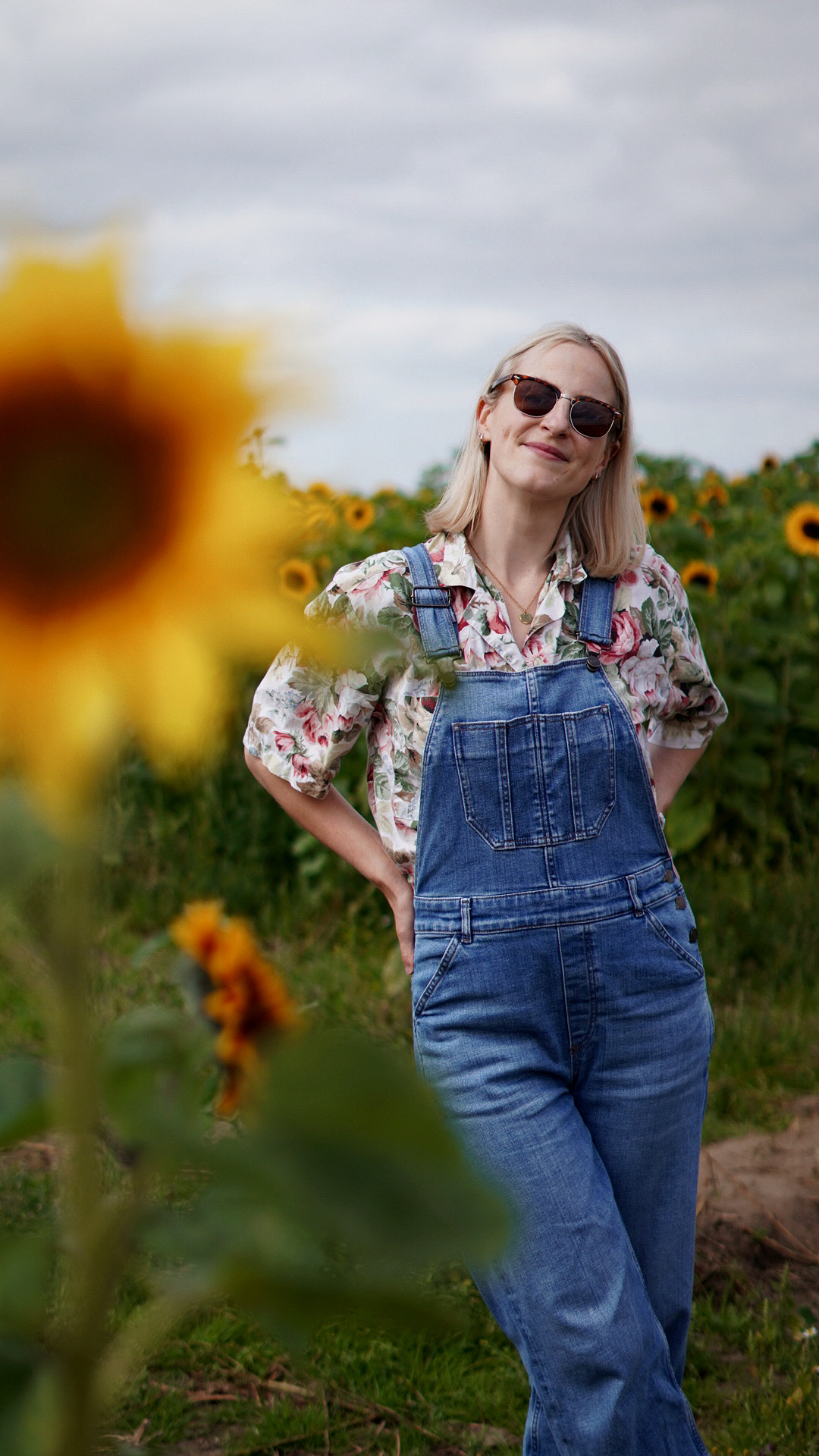 Amy is standing in a field of sunflowers, she is wearing a floral shirt and denim dungarees and looking to camera smiling