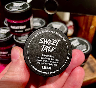 A white hand holding a glass cylindrical jar containing neon orange lip scrub with a black lid  that has a black circular lid that says sweet talk lip scrub lush in white font on a bright background