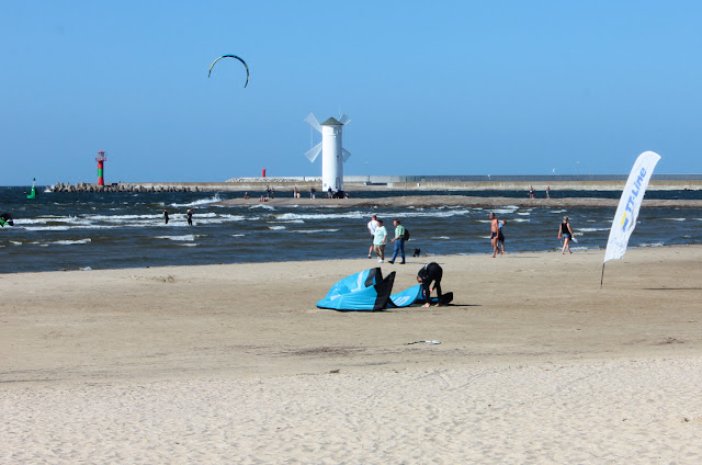 Na plaży w Świnoujściu z widokiem na falochron i zdobiacy go biały znak nawigacyjny, w kształcie wiatraka. Przy falochronie biorę fale i wiatr kitesurferzy.