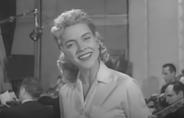 Blonde Ella Mae Morse wearing a white blouse, smiling while looking into the TV camera, with an orchestra seated behind her and a large microphone aimed at the orchestra.