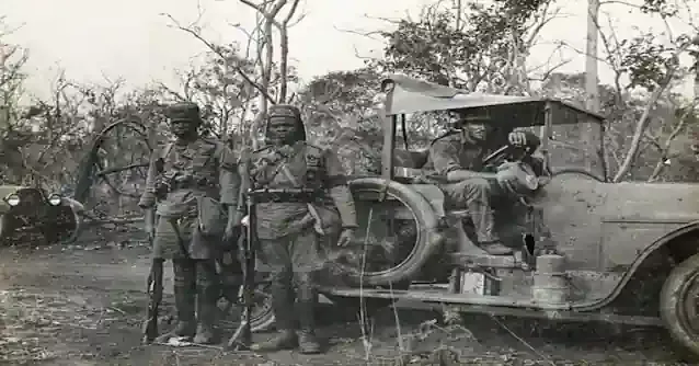Two soldiers from the King's African Rifles (KAR) standing beside a military vehicle with personnel, possibly of British origin