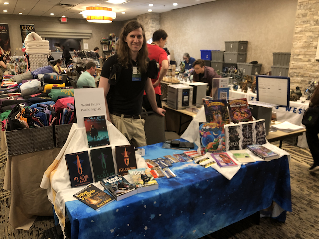 This photo by Pascal Gephardt shows the Weird Sisters Publishing Dealers Table at DemiCon 34. Tyrell Gephardt stands behind the table. On the left-hand side, from top to bottom of the display, are copies of Dora Furlong’s “One of Our Own,” then Lynette M. Burrows’ “My Soul to Keep,” “If I Should Die,” and “Fellowship.” On the table level are Jan S. Gephardt’s “The Other Side of Fear,” “What’s Bred in the Bone,” and “A Bone to Pick.” In the middle of the table, we attached badge ribbons to bookmarks and business cards with information about the books the quotes come from. To get a badge ribbon, table visitors also had to take the attached information. On the right side of the table, from top to bottom, are Randal Spangler’s hardbound, fully illustrated children’s books, “D is for Draglings™” and “The Draglings™’ Bedtime Story.” On the next level are “The Draglings™ Coloring Book” and the three volumes of Karin Rita Gastreich’s “Silver Web Trilogy,” “Eolyn,” “Sword of Shadows,” and “Daughter of Aithne.” On the table level are G. S. Norwood’s “Deep Ellum Duet” and M. C. Chambers’ “Midsummer Storm” and “Shapers’ Veil.” Tablecloth design is “Nebula 2,” ©2021 by Chaz Kemp.