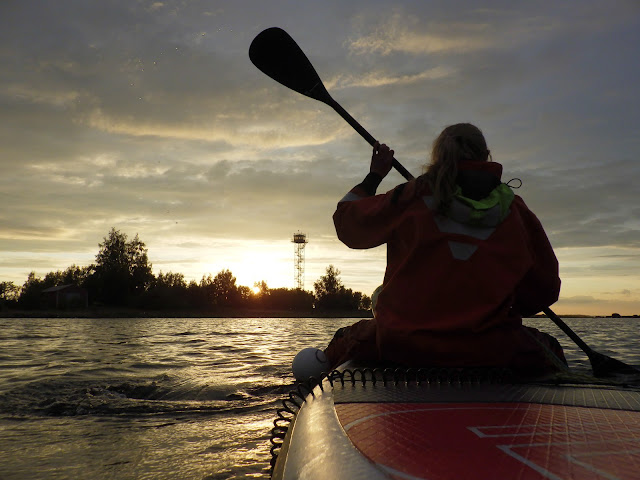 Pelastautumispukuinen henkilö melomassa SUP-laudalla kohti saarta auringonlaskussa