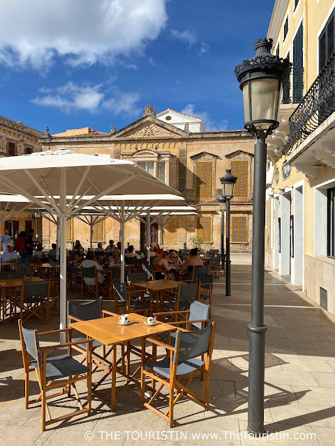 A cafe on a square lined by sandstone period properties with the focus on two white espresso cups on an empty wooden bistro table with four grey director chairs next to a period-style streetlight, in front of guests sitting on wooden and light grey director chairs at wooden bistro tables under large white sun umbrellas.