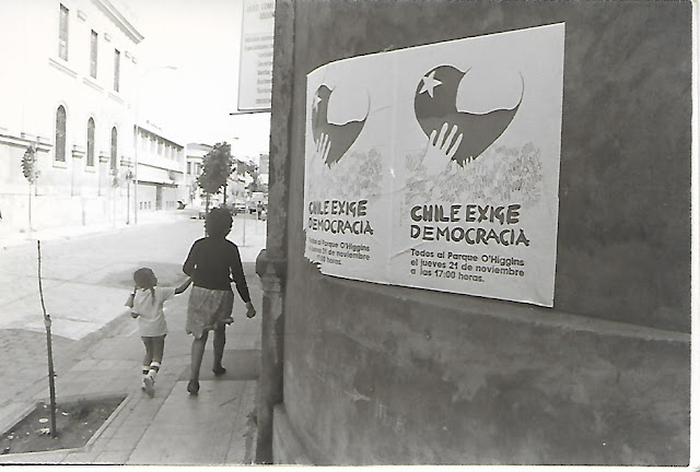 A fotografia em preto e branco mostra uma mulher e uma criança de costas andando na calçada, ao lado do muro da casa pela qual passam está colado um cartaz com uma chamada para uma manifestação em defesa da democracia chilena.
