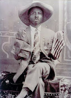 Prophet Noble Drew Ali seated wearing a sombrero hat holding the US and Moorish flags in his right and left hands.