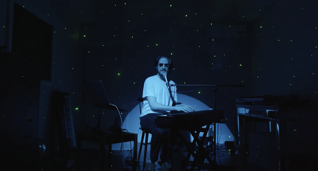 A still from Inside. Bo Burnham sits in darkness with a spotlight on him. He is wearing sunglasses and playing a keyboard.