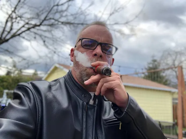 2/3 Man in his late 40s smoking a cigar wearing a black leather jacket is glasses on