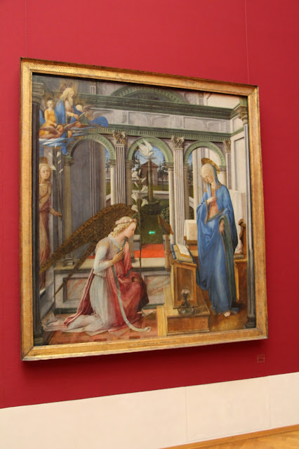 Renaissance annunciation scene. Fanciful architectural elements. A feminine angel Gabriel kneeling (long hair, wreath around head) before the Virgin Mary. Gabriel is in red and white. Mary is in Blue and white and is standing. A dove looks on. Gabriel looks at the floor and holds a tall lily.