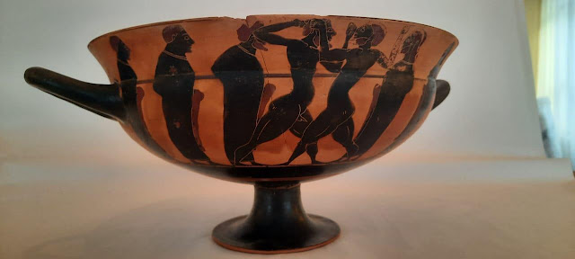 Μελανόμορφη κύλικα τύπου Σιάννων με παράσταση πυγμάχων παρουσία κριτών. Στον τόντο απεικονίζεται κόκορας. Γύρω στα μέσα του 6ου αι. π.Χ.​
