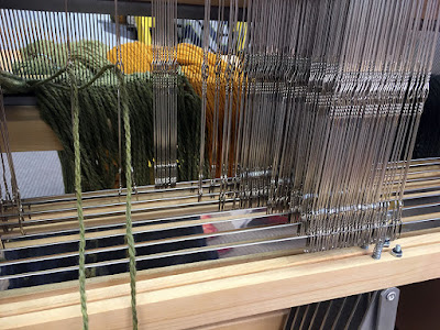 Metal heddles seen from the back of a loom, with two strand of light green wool yarn threaded at middle left, and scores of empty heddles at right. Light green and marigold warp yarn is visible through the heddles, hanging through the reed.