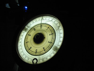 Photo of a brightly-lit circular white gauge against a nearly-black background. The gauge is round with three concentric rings of numbers. The innermost ring runs from 0 to 9 with tick marks for fifths in between each numeral. The next ring out has extremely fine numerical divisions that can't be read on the photo. The outermost ring runs from 0 to 360 in increments of 10, each further divided by ticket marks.