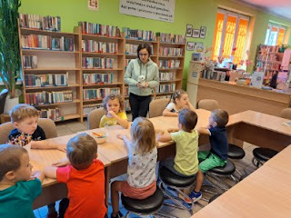 Zdjęcie ukazuje grupę przedszkolaków siedzącą przy jasnobeżowych stolikach. Pani bibliotekarka stoi na tle regałów z książkami.