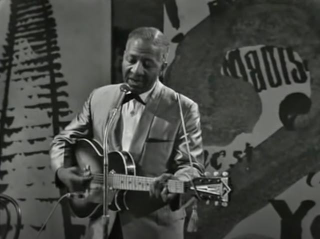 Screenshot from a 1963 video of Lonnie Johnson playing his guitar and singing. He's wearing a suit and stands in front of a collage background on the stage.