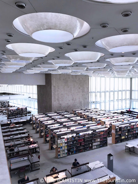 Nine people sit randomly at desks in a light-filled airy large concrete hall with several rows of white bookshelves under huge round white ceiling lights.