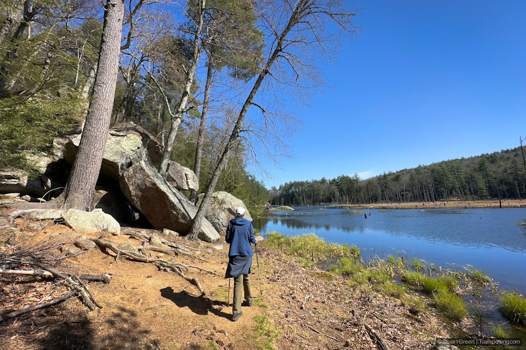 A hiker walking alongside a blue pond, with large boulders towering overhead.