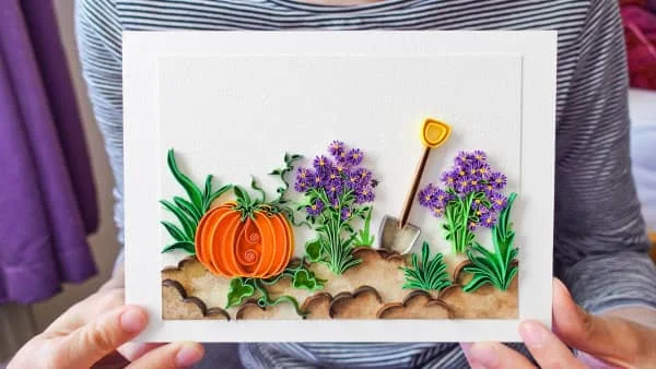 hands holding quilled scene of pumpkin growing in garden with flowers and shovel nearby