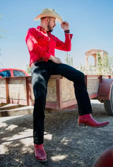 5/11 sitting slightly Superior POV with a light straw cowboy hat tipping for with hand wearing bright hot pink dress shirt dark blue jeans and matching leather cowboy crocodile boots with the hot flamingo pointing to the side sitting outside on a ranch fence