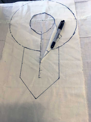 A rectangle of near-white fabric with torn edges, a small circle cut from the center, and a larger circle marked in black sharpie, with a line connecting the two circles' lowest points and continuing downward, with hashmarks at regular intervals on the descender. A broad, down-pointing border is marked around the descender, starting at the large circle, in black sharpie. The sharpie is restin gon a folded-back corner of the collar design, showing the second layer of near-white fabric beneath.