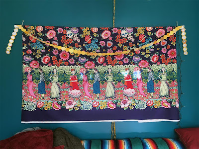 A long chain of yellow hexagons, draped over a hanging panel of purple, multicolored floral fabric with a border of Frida Kahlo figures, against a bright teal wall.