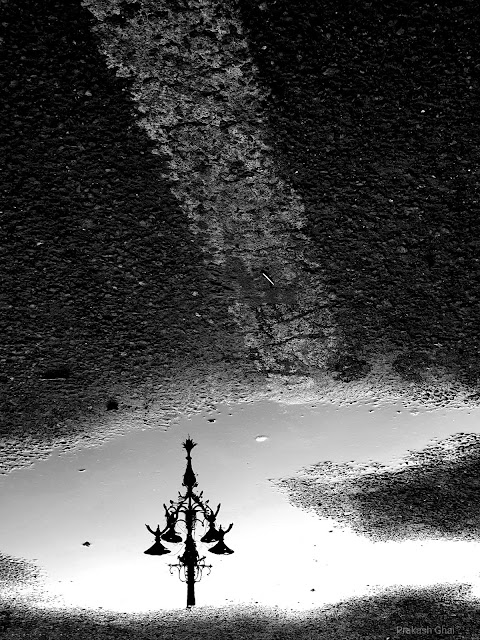 A Black and White Minimalist Photograph of the Reflection of a Lamp in a puddle of Water, at Panch Batti MI Road, Jaipur