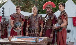 Four Roman officers, of various levels of seniority, around a map table, in front of a large white tent.