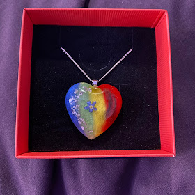 A silver chained necklace with a large pendant in its box. The pendant is heart-shaped clear acrylic. In the background of the pendant is a rainbow with vertical stripes. In the foreground on the left are some grey speckles, in the middle is a dried forget-me-not (a small blue flower), and on the right is a lock of black fur.