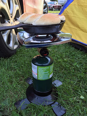 A single-burner propane camping stove, with a small cast-iron skillet on top and a split hot dog bun resting on top of the contents, set up in grass in a corner formed by a car wheel and a bright yellow-and-blue tent.