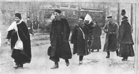 Black and white photo of seven African men walking down a street. They are all wearing turbans and wearing a thick black winter fur coat. Behind them passes by a tram.