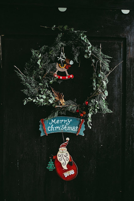 Corona navideña colgada en una puerta, de ramas de abeto y eucalipto, con reno y Santa Claus colgados de la corona y cartel de Merry Christmas