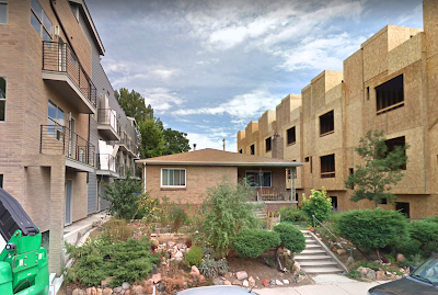 1-story house at center on a slight rise with a 3-story apartment building at left and another 3-story apartment building under construction on the right.