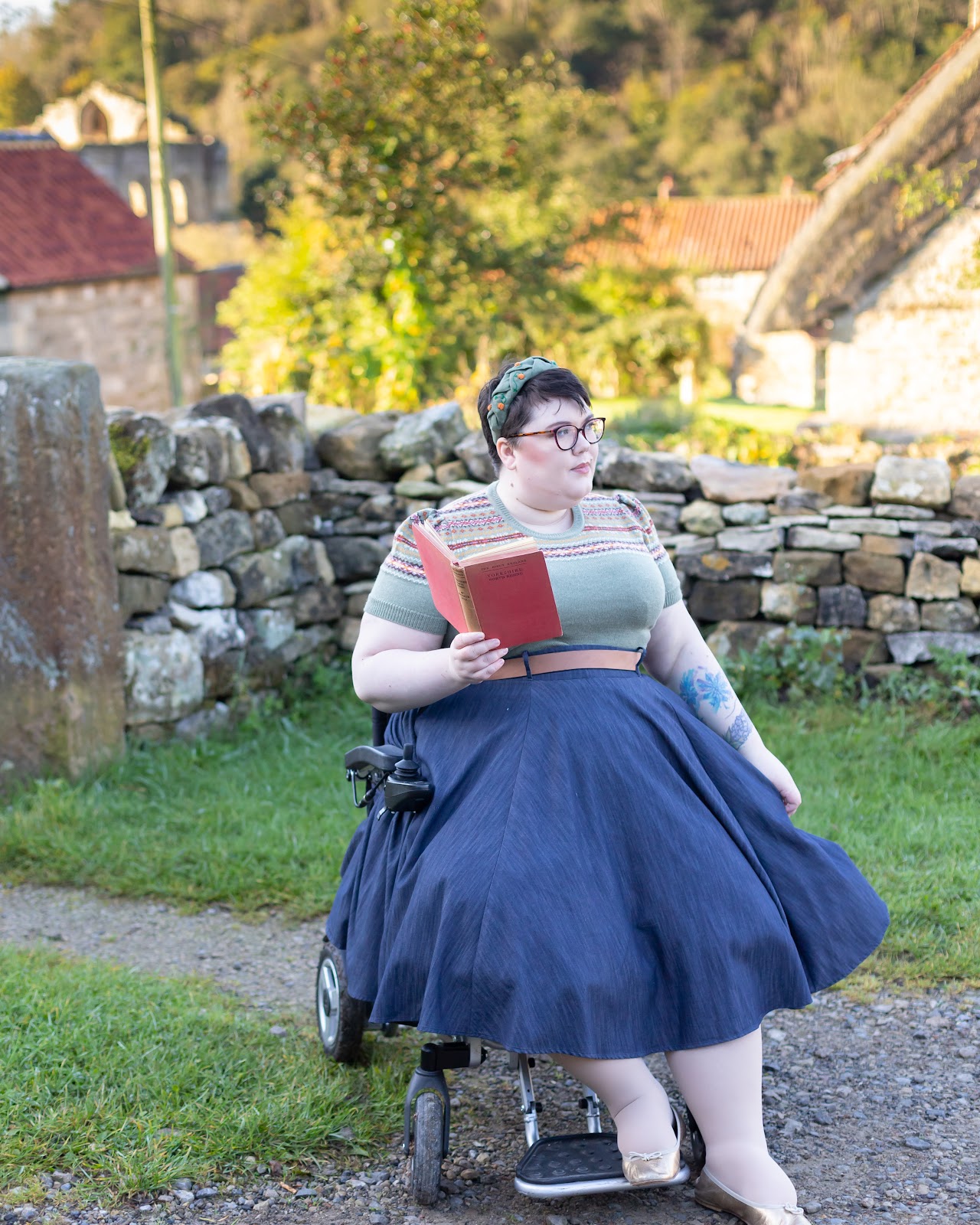 A plus size white person sits in an electric wheelchair wearing a 1940s cottagecore outfit and holding a red book