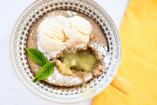 A microwave apple sponge pudding in a large mug topped with vanilla ice cream and a sprig of mint