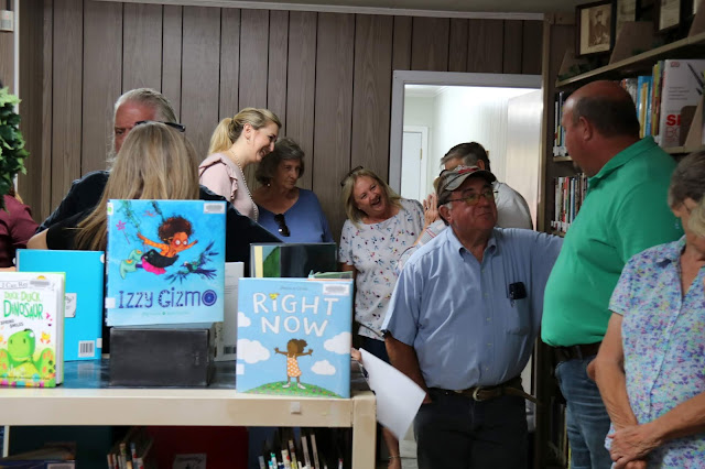 A large group of people are standing around talking. They are crammed into a library and children's books are arranged on the tops of bookshelves.