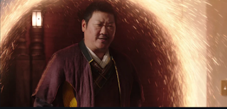 Wong, a heavyset Asian man in his middle years, dressed in robes. He's stepping through a portal of spinning orange light in the air.