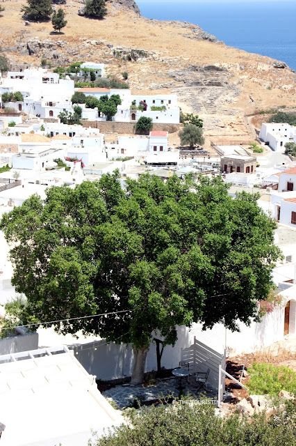 A round table with two chairs in the shade of a green tree with a white village in the background.