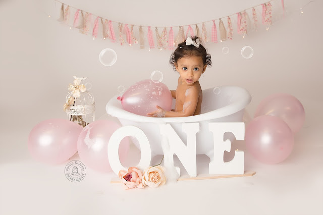 Little girl sits in a small white bathtub surrounded by pale pink balloons, soap bubbles, and flowers. Three white wooden signs with the letters “O”, “N”, “E” lean against the bathtub just below her