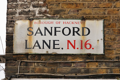 Photograph of a street sign, black and red letters on white, saying 'BOROUGH OF HACKNEY SANFORD LANE, N.16.'